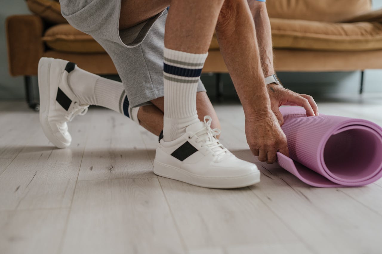 Senior man in sneakers rolling a purple yoga mat on a wooden floor indoors.